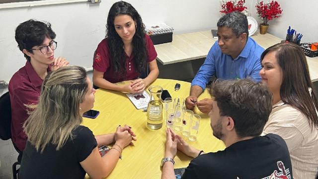 Helder Eterno e Fabiola Gomes estão à direita, sentados em volta de uma mesa redonda, conversando com quatro estudantes