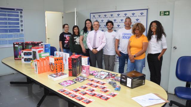 Oito pessoas em pé, posando para a foto; na mesa, estão expostos os itens doados pela Receita Federal à Dirco