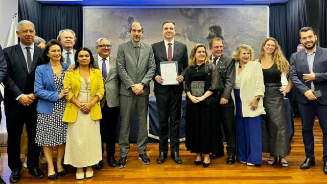 Imagem de 12 pessoas em pé; ao centro, o presidente do Senado exibe a homenagem recebida