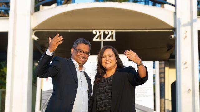 Foto de Hélder Eterno e Fabíola Gomes em frente à entrada da UFU no Campus Santa Mônica; ambos estão em pé, sorridentes e fazendo gesto com uma mão aberta