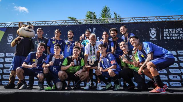 Imagem de uma equipe masculina reunida no primeiro lugar de um pódio, exibindo medalhas