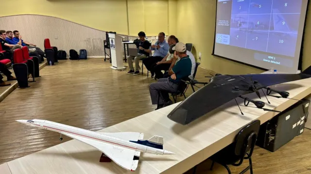No primeiro plano, uma mesa na diagonal com dois mini protótipos de aviões, e no segundo plano da imagem, alguns palestrantes do evento sentado em cadeiras para o momento de roda de conversa