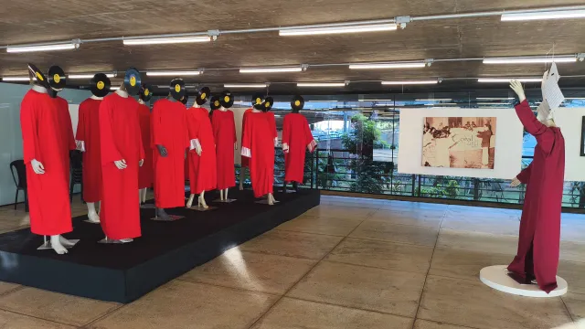 Exposição artística com manequins de vestes vermelhas. À esquerda, sobre um tablado baixo, um grupo de manequins usa discos de vinil no lugar das cabeças. À direita, um manequim isolado em túnica escura estende os braços como um regente em frente a uma imagem na parede.