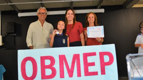 Há um grupo de quatro pessoas atrás de um banner com a palavra 'OBMEP'. Uma dessas pessoas é uma menina que segura uma medalha